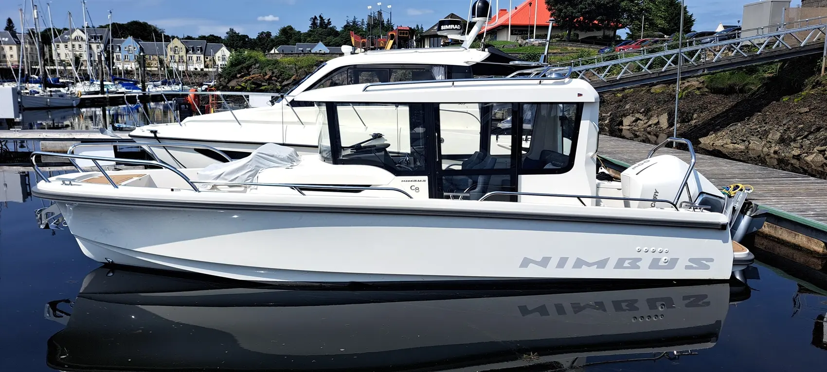 A Nimbus C8 motorboat docked in a marina, reflecting in calm waters, with homes and other boats in the background.